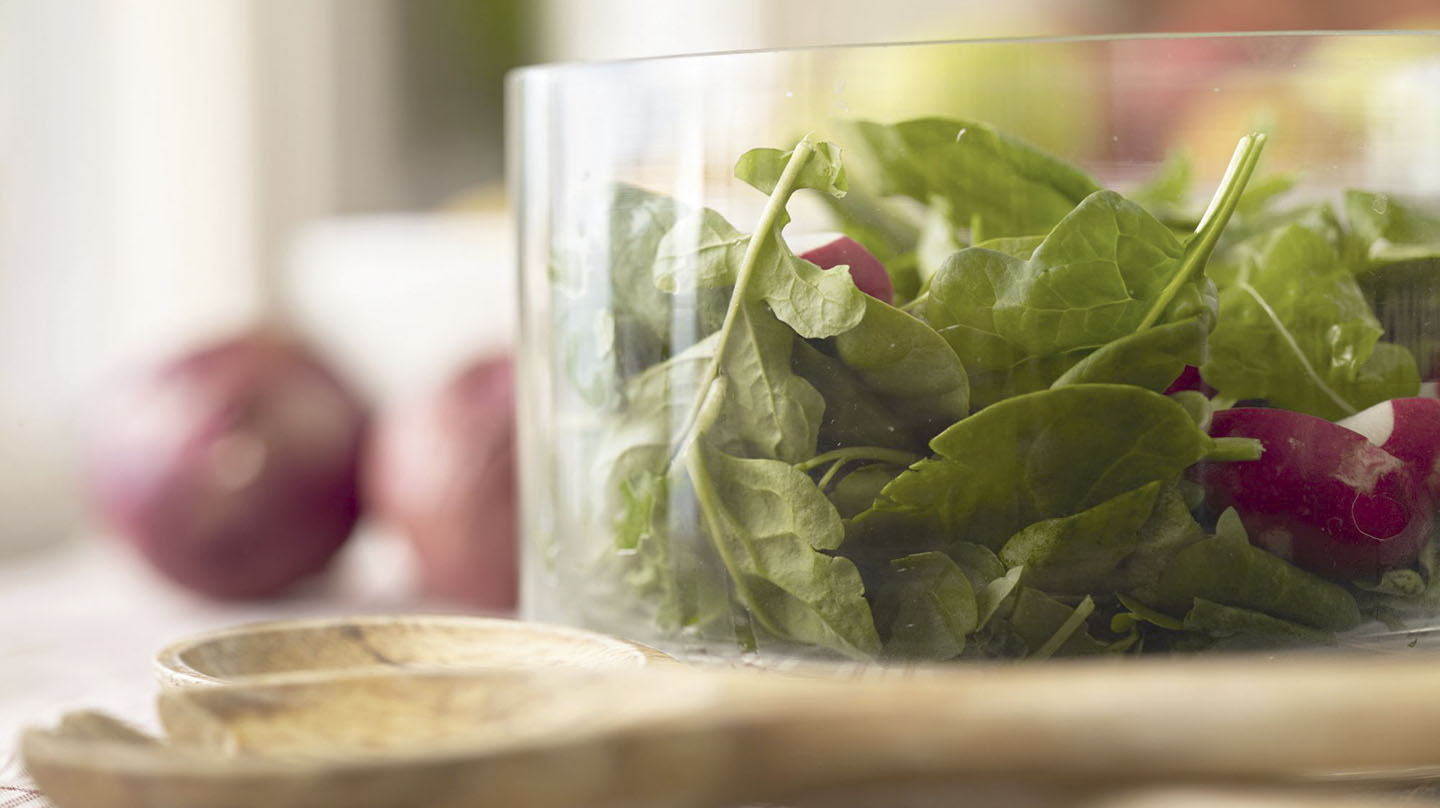 Lebensmittel für konstanten Blutzucker: Glasschüssel mit Salatblättern und Radieschenstücken, davor liegt ein Salatbesteck aus Holz, im Hintergrund zwei rote Zwiebeln.